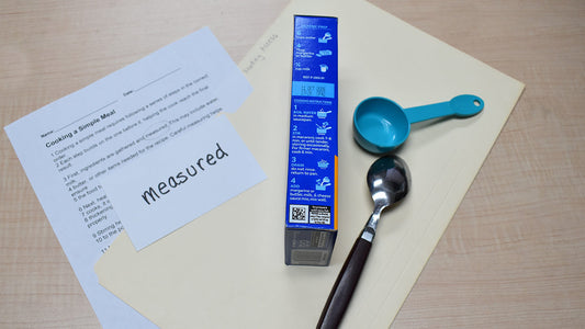 Folder labeled Reading Access with a reading passage titled Cooking a Simple Meal partially visible, a handwritten vocabulary card on top, and a macaroni and cheese box with cooking instructions, spoon, and plastic measuring cup