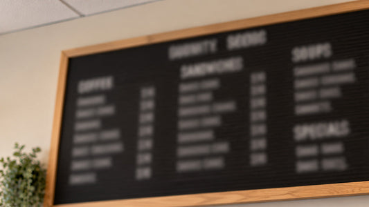Out-of-focus menu board with white lettering in a wooden frame, surrounded by a blurred wall, ceiling, and plant.