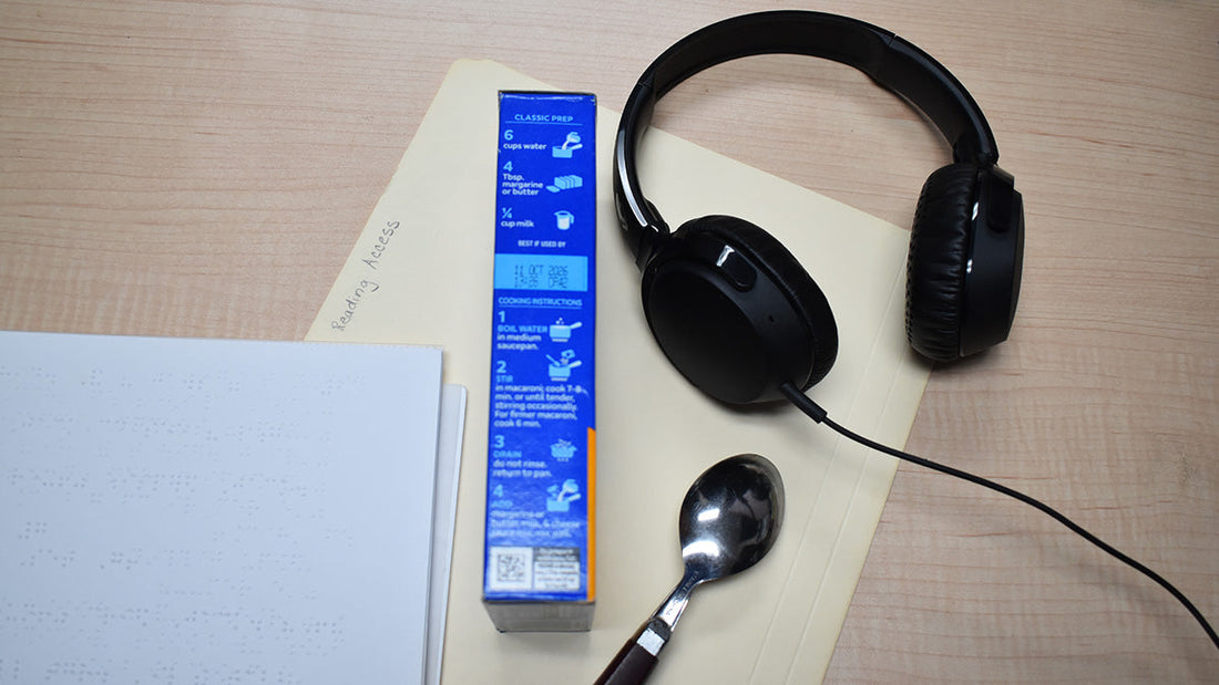 Folder labeled Reading Access with braille pages partially visible, a macaroni and cheese box with cooking instructions, a spoon, and headphones arranged on the folder.
