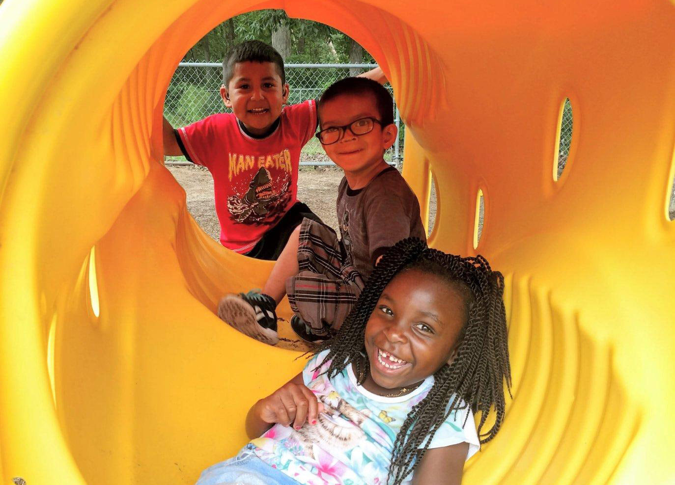 Three young children play in a yellow playground tunnel.  Two children have hearing aids and one has glasses.  The children are looking happy and laughing.